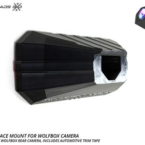 Può includere: Un supporto di superficie nero, angolare e sfaccettato progettato per una telecamera posteriore Wolfbox. Il supporto presenta un interno cavo, una finitura esterna testurizzata e include nastro adesivo automobilistico. Il testo CROSSROADS 360 è visibile.