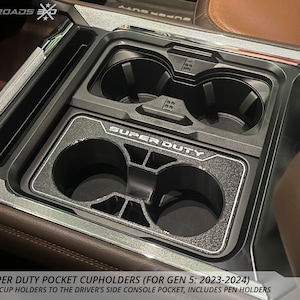 May include: A black Ford Super Duty pocket cupholder with the text "SUPER DUTY" visible. The cupholder includes pen holders and is designed for the driver's side console pocket. The product is made in the USA.