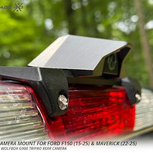 以下が含まれることがあります： Ford F150 (15-25) および Maverick (22-25) 車両用の黒い Wolfbox カメラマウント。マウントは銀色のトップです。画像には赤いテールライトと「MADE IN THE USA」の文字も表示されています。