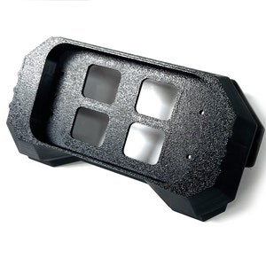 May include: A black, textured plastic component with four square openings and a geometric design. The object has a rectangular shape with angled sides and appears to be a housing or mounting bracket. The surface has a speckled texture.
