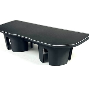 May include: A black car console made of plastic, featuring a textured surface and a white trim. It has two cup holder spaces. The console is designed for a vehicle and is shown against a white backdrop.
