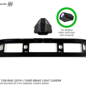 May include: A black plastic third brake light camera mount for 2019+ RAM trucks, featuring a central housing for Wolfbox G900 and G930 style cameras. The set includes a main mounting bracket and a separate camera cover piece with text labels.