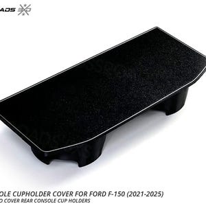 May include: Black rear console cupholder cover for Ford F-150 (2021-2025). The cover has a textured surface with a white trim. The product is made in the USA. The text on the image reads: REAR CONSOLE CUPHOLDER COVER FOR FORD F-150 (2021-2025) PRESS-IN CAP TO COVER REAR CONSOLE CUP HOLDERS.