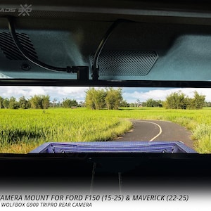 以下が含まれることがあります： 緑の野原を通る道路を示す車のバックミラー。ミラーは車両に取り付けられており、下部に「WOLFBOX CAMERA MOUNT FOR FORD F150 (15-25) & MAVERICK (22-25)」の文字があります。アメリカ製。