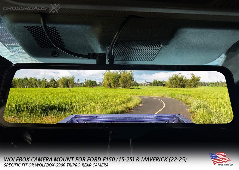 以下が含まれることがあります： 緑の野原を通る道路を示す車のバックミラー。ミラーは車両に取り付けられており、下部に「WOLFBOX CAMERA MOUNT FOR FORD F150 (15-25) & MAVERICK (22-25)」の文字があります。アメリカ製。