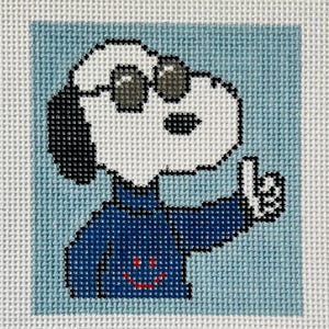 May include: Needlepoint canvas featuring a cartoon dog wearing sunglasses and a blue outfit, giving a thumbs-up gesture. The dog has a white face, black ears, and a red smiling mouth. The background is light blue.