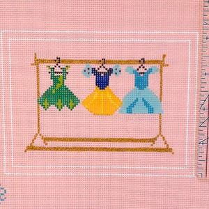 Puede incluir: Dise&ntilde;o de punto de cruz con tres vestidos colgados de un perchero dorado. Los vestidos son verdes, amarillos y azules, cada uno con detalles &uacute;nicos. El fondo es una tela rosa claro con un borde blanco y una regla en el lado derecho.