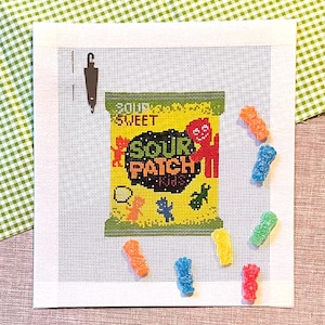 May include: Needlepoint canvas featuring a Sour Patch Kids candy bag design. The bag is yellow and green with the words "Sour Patch Kids" in a bold font. Scattered around the canvas are colorful gummy candies in various shapes and colors.