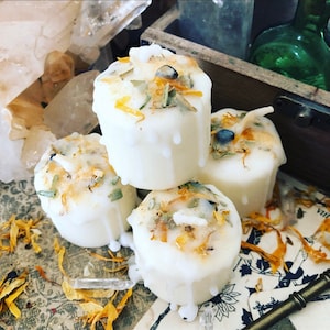 May include: Four white votive candles with dried herbs and flower petals on top. The candles have a dripped wax design and are surrounded by dried petals and crystals. The candles are a part of a spiritual or decorative theme.