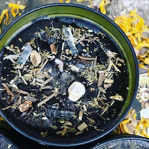 May include: A black candle in a round, green-rimmed container. The candle is decorated with a small white skull, crystals, and dried herbs. The candle is surrounded by dried yellow flower petals.
