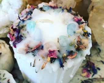 May include: A white candle with a melted wax exterior, adorned with light blue crystals and dried flower petals in shades of pink, blue, and yellow. The candle has a single wick and is surrounded by other candles.