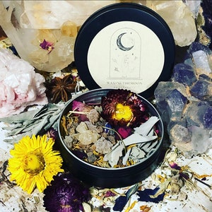 Puede incluir: Una lata de metal negro con tapa crema que presenta un diseño de media luna y el texto "WAXINGTHEMOON apothecary". La lata abierta revela flores secas, hierbas y cristales. Cristales y flores rodean la lata.