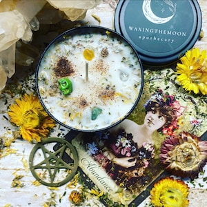 May include: A candle in a black tin with a white wax base, adorned with herbs, crystals, and a green skull. The tin lid displays the text "waxingthemoon apothecary". Dried flowers and a vintage illustration with the word "Abundance" surround the candle.