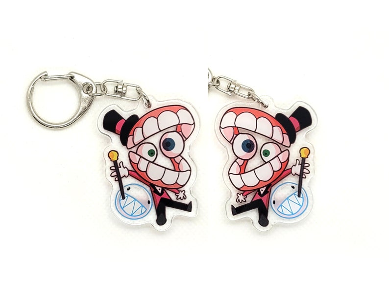 May include: Two red and white cartoon character keychains with a toothy grin, wearing a top hat and holding a stick. The characters are holding a blue and white circle with a jagged edge.