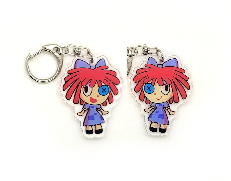 May include: A red-haired doll with blue eyes and a blue and white dress is attached to a silver keychain. The doll has a stitched X over one eye.