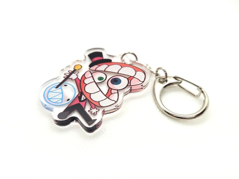 May include: A clear acrylic keychain featuring a cartoon character with a red and white hat, a large mouth with white teeth, and one blue eye. The character is holding a stick with a star on the end. The keychain is attached to a silver metal ring.