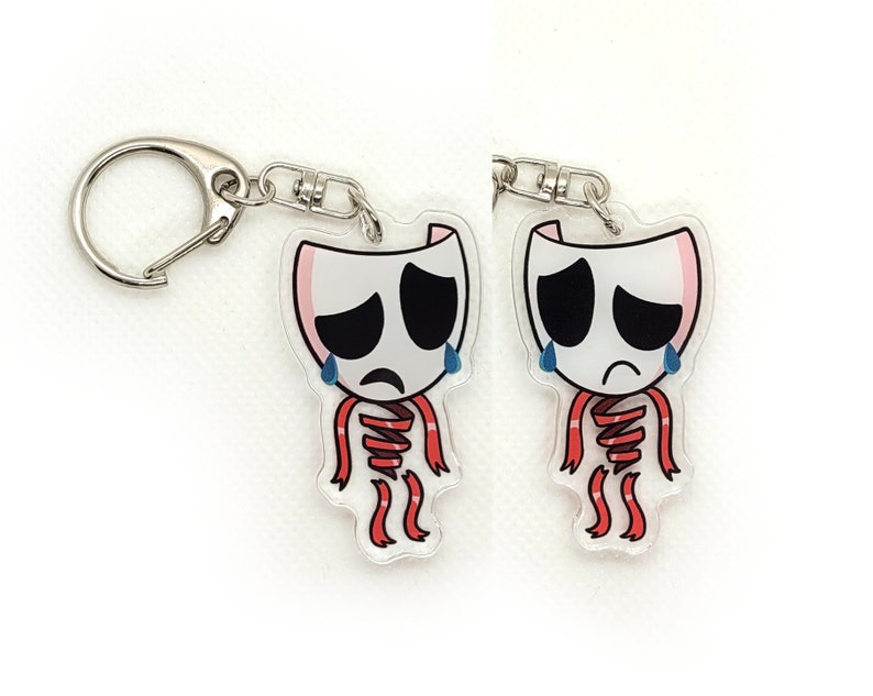 May include: Two clear acrylic keychains with a sad face and red ribbon design. The keychains are attached to silver key rings.