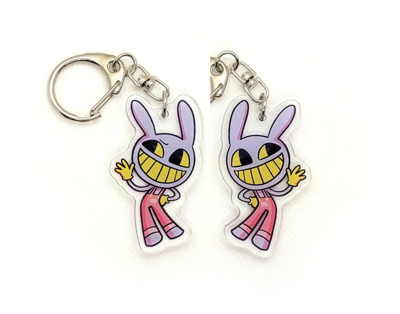 May include: Two purple and white cartoon bunny keychains with a wide grin and yellow teeth. The bunny is wearing pink overalls and is waving with one hand. The keychains are attached to silver key rings.