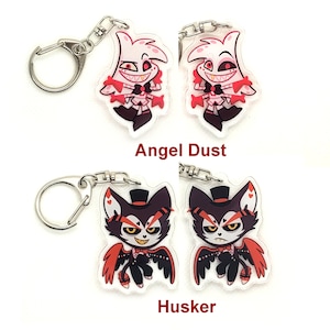 Puede incluir: Dos llaveros con personajes de la serie animada Hazbin Hotel. El primer llavero presenta a Angel Dust, un demonio rosa y blanco con cuernos rojos y una corbata de mo&ntilde;o roja. El segundo llavero presenta a Husker, un demonio negro y rojo con un sombrero de copa y alas rojas. Ambos llaveros est&aacute;n sobre un fondo blanco.