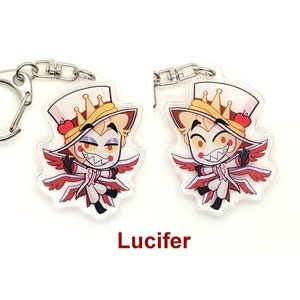 Puede incluir: Un llavero con un personaje de dibujos animados con alas rojas, un sombrero blanco con una corona y una manzana roja. El personaje tiene el pelo amarillo, los ojos rojos y una sonrisa traviesa. El texto "Lucifer" est&aacute; impreso debajo de la imagen.
