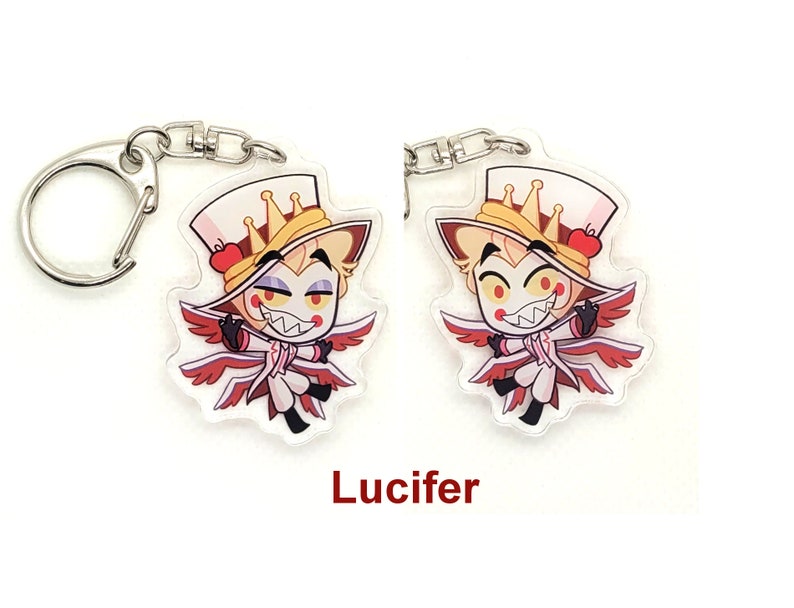 Puede incluir: Un llavero con un personaje de dibujos animados con alas rojas, un sombrero blanco con una corona y una manzana roja. El personaje tiene el pelo amarillo, los ojos rojos y una sonrisa traviesa. El texto "Lucifer" est&aacute; impreso debajo de la imagen.