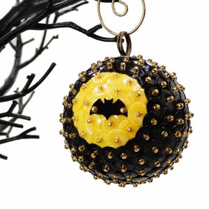 May include: A black and yellow Christmas ornament with a bat silhouette in the center. The ornament is made of sequins and beads.