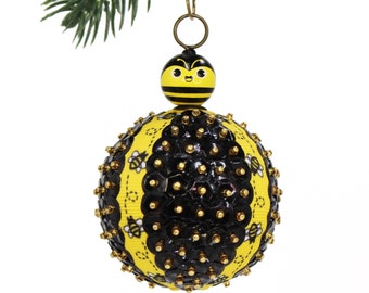 Puede incluir: Un adorno navideño redondo de color amarillo y negro con temática de abejas con una cara de abeja en la parte superior. El adorno está cubierto de lentejuelas negras y tiene una cuerda dorada para colgar.