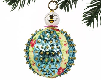 Puede incluir: Un adorno navideño de color turquesa y amarillo con un diseño de abeja. El adorno está cubierto de lentejuelas y tiene un patrón floral.