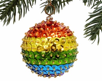 May include: A round Christmas ornament with a rainbow color scheme, hanging from a gold-colored chain. The ornament is covered in small, shiny sequins. The ornament is hanging from a green pine branch.