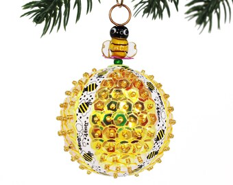 Puede incluir: Un adorno navideño dorado y blanco con forma de esfera con un diseño de abeja. El adorno está cubierto de lentejuelas y tiene una abeja negra y amarilla en la parte superior.