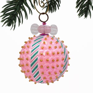 May include: A spherical Christmas bauble featuring alternating vertical panels of pink sequins with gold beads and white stripes with green glitter accents. The top is adorned with a clear glass bead shaped like a sweet and a metal hanging loop.