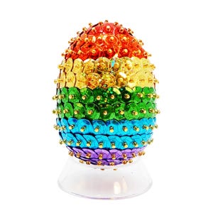May include: A rainbow-colored Easter egg decorated with sequins. The egg is on a clear stand.