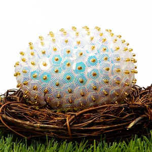 May include: A white iridescent Easter egg decorated with gold sequins and beads sits in a brown twig nest on a bed of green grass.