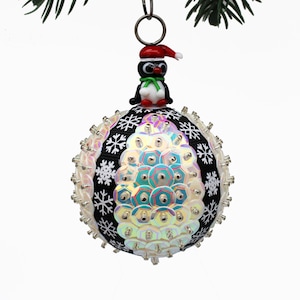 May include: A festive Christmas bauble featuring a penguin in a Santa hat. The round decoration is white with iridescent sequins and black snowflake patterns. A silver loop is at the top.