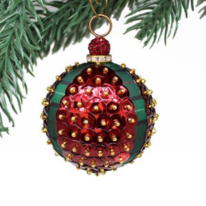 May include: A spherical Christmas ornament featuring a green and black plaid fabric base. The surface is decorated with a central teardrop-shaped panel of shiny red sequins, accented with gold beads. A red sequined cap sits at the top with a metal ring.