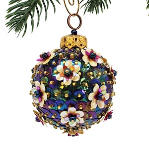 Peut inclure: Une décoration de Noël avec un motif floral. L'ornement est une sphère recouverte de paillettes irisées et ornée de petites fleurs crème avec des accents roses et bleus. Il est suspendu à un capuchon en métal doré.
