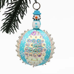 May include: A festive Christmas bauble with a snowman and a blue and white design. The round ornament has a light blue base, decorated with sequins and snowflakes. A small snowman figure with a blue hat sits on top of the bauble.