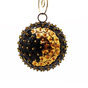 May include: A spherical holiday ornament covered in overlapping black and gold sequins. The ornament features a gold wire loop at the top for hanging. Each sequin is secured with a small gold pin, creating a textured, reflective surface.