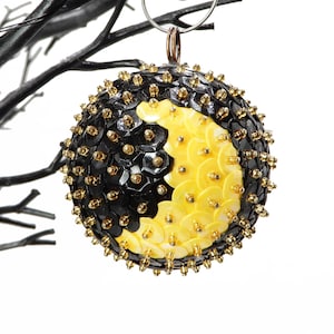 May include: A round Christmas ornament with a black and yellow design. The ornament is covered in sequins and small gold beads. It hangs from a silver loop and is set against a white background.