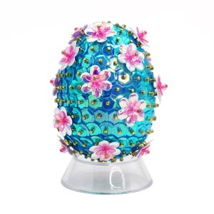 May include: A blue Easter egg covered in iridescent sequins and decorated with pink flowers. The egg is sitting on a clear plastic stand.