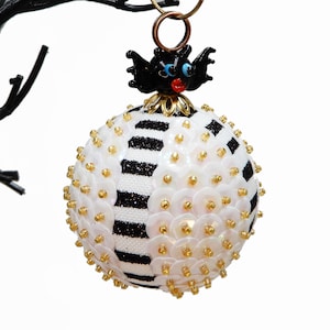 May include: Black and white striped Christmas ornament with white sequins and gold beads. The ornament has a black bat with red lips and blue eyes on top.