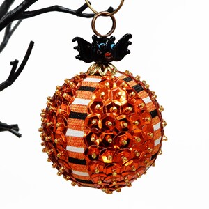 May include: Orange and black Halloween ornament with a bat on top. The ornament is covered in sequins and has a striped pattern.