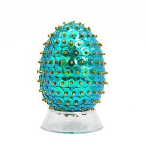 May include: A teal blue Easter egg covered in sequins with gold accents on a clear plastic stand.