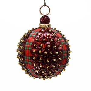 May include: A spherical holiday ornament featuring alternating panels of red and green plaid fabric and sections covered in deep red sequins. Each sequin is secured with a small gold bead. The top includes a gold band and a red beaded cap.