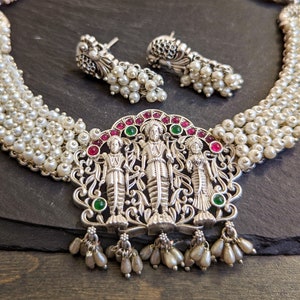 May include: A silver necklace with a pendant featuring three figures and green and pink gemstones. The necklace is made of multiple strands of white pearls.