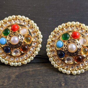 May include: A pair of gold-tone earrings with a circular design. The earrings are adorned with a variety of colorful gemstones, including red, green, blue, and orange. The gemstones are surrounded by a ring of smaller white stones.