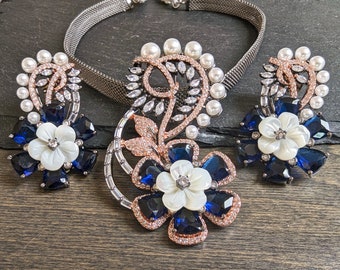 May include: A silver necklace with a blue and white flower pendant and matching earrings. The pendant features a pearl and crystal accents. The earrings are also adorned with blue and white flowers.