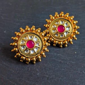 May include: A pair of gold-colored stud earrings. Each earring features a central, round, magenta-colored stone surrounded by clear stones and a gold-colored beaded border. The earrings have a sunburst design.