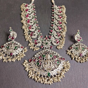 May include: A silver necklace and earrings set with a traditional Indian design. The necklace features a pendant with a goddess figure, surrounded by intricate floral patterns and red and green gemstones. The earrings are similarly designed with a smaller pendant and pearl drops.