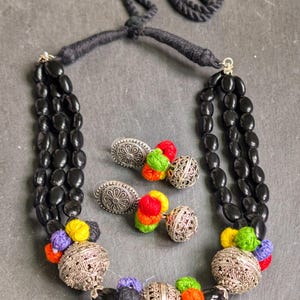 May include: A black beaded necklace and matching earrings set. The necklace features three strands of black beads, silver-colored metal accents, and colorful yarn embellishments. The earrings have silver-colored metal and colorful yarn accents.
